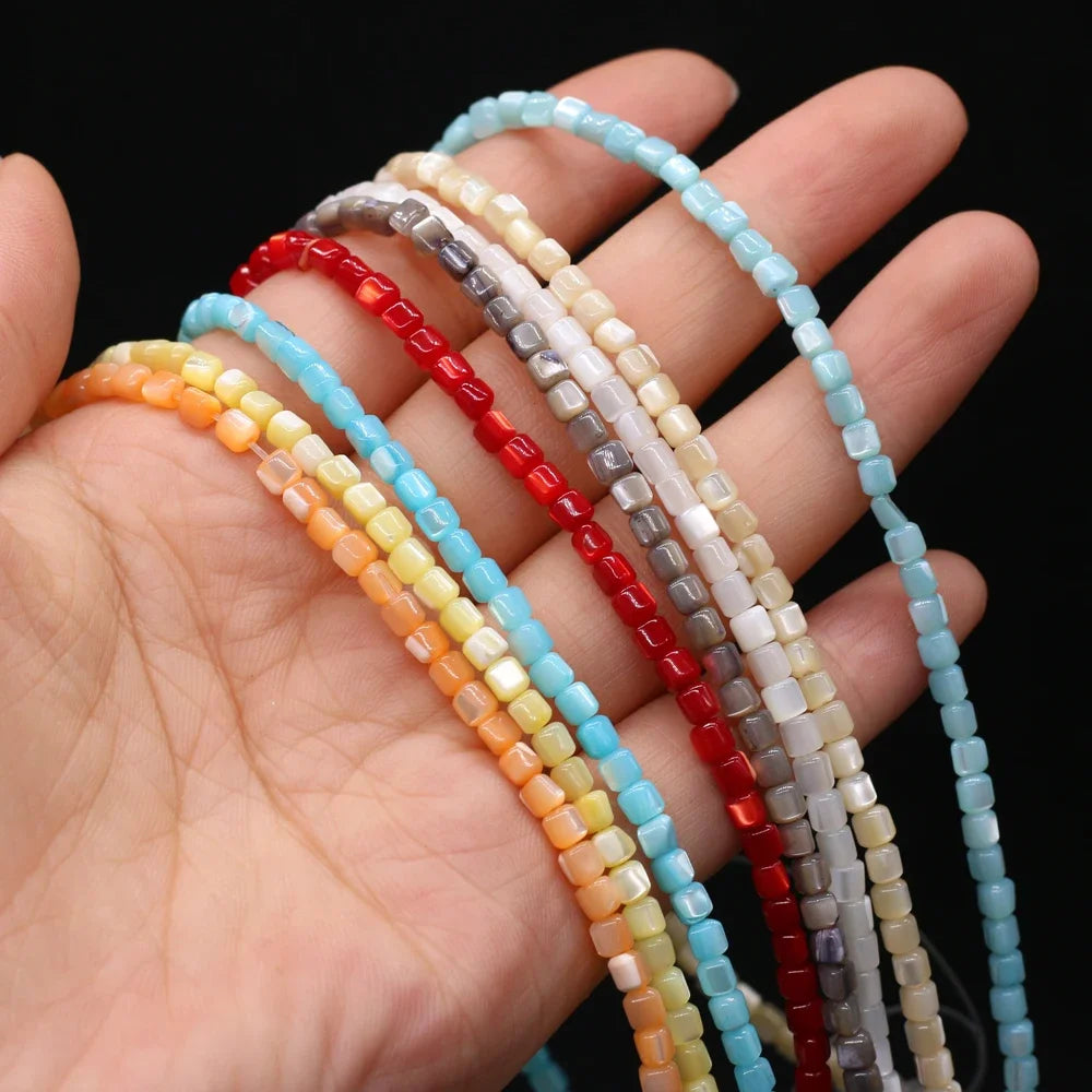 Multiple strands of small square shell beads in assorted colors, including light blue, red, yellow, orange, gray, and cream, draped over a person's hand against a black background.