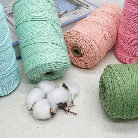 Four large spools of twisted cotton twine in mint green, peach pink, light blue, and sage green, arranged on a light surface with a few cotton bolls in front.