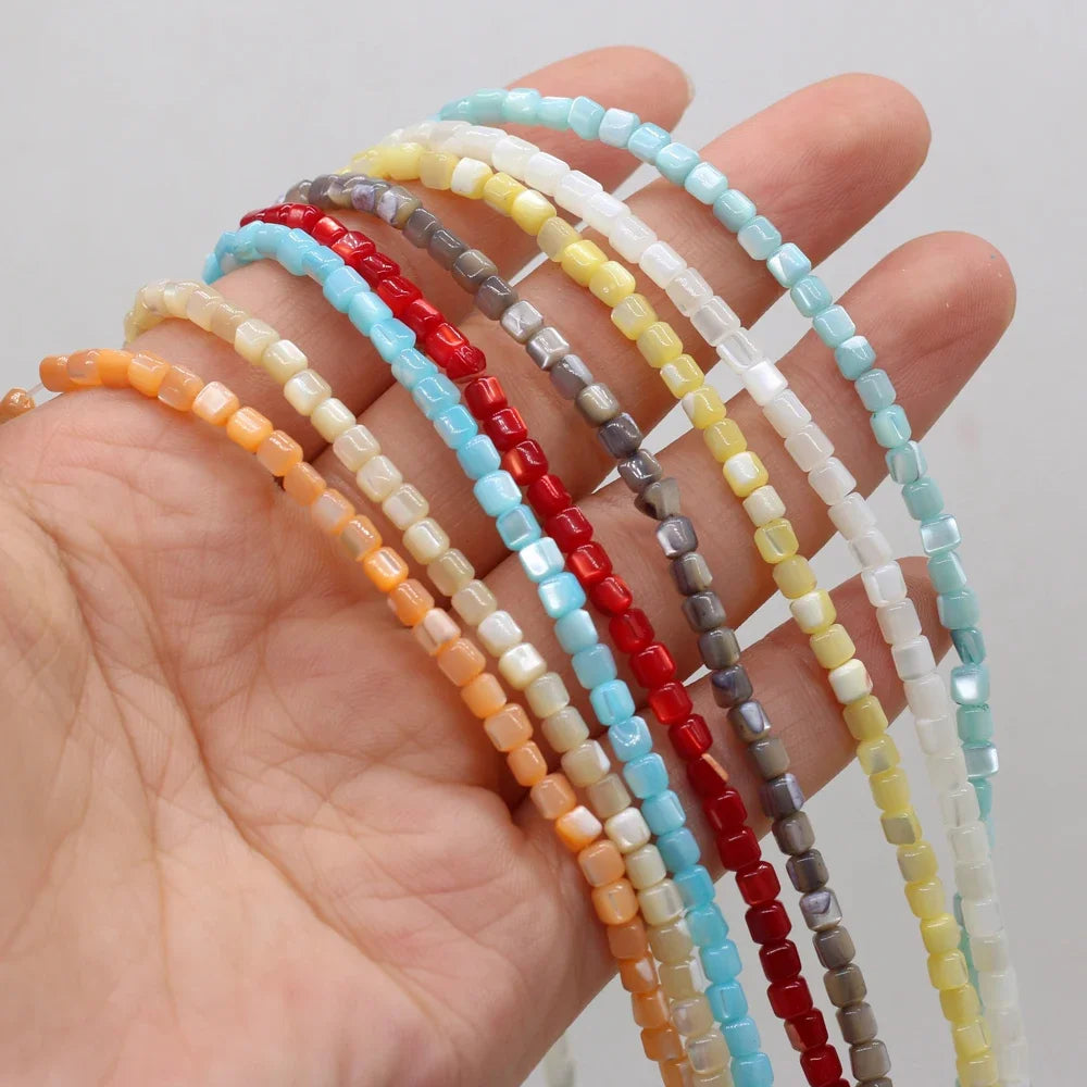 A hand holding several strands of small rectangular shell beads in assorted colors, including light blue, red, gray, yellow, white, and orange, against a plain light background.