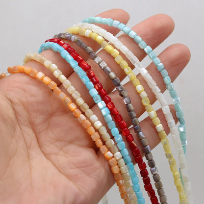 A hand holding several strands of small rectangular shell beads in assorted colors, including light blue, red, gray, yellow, white, and orange, against a plain light background.
