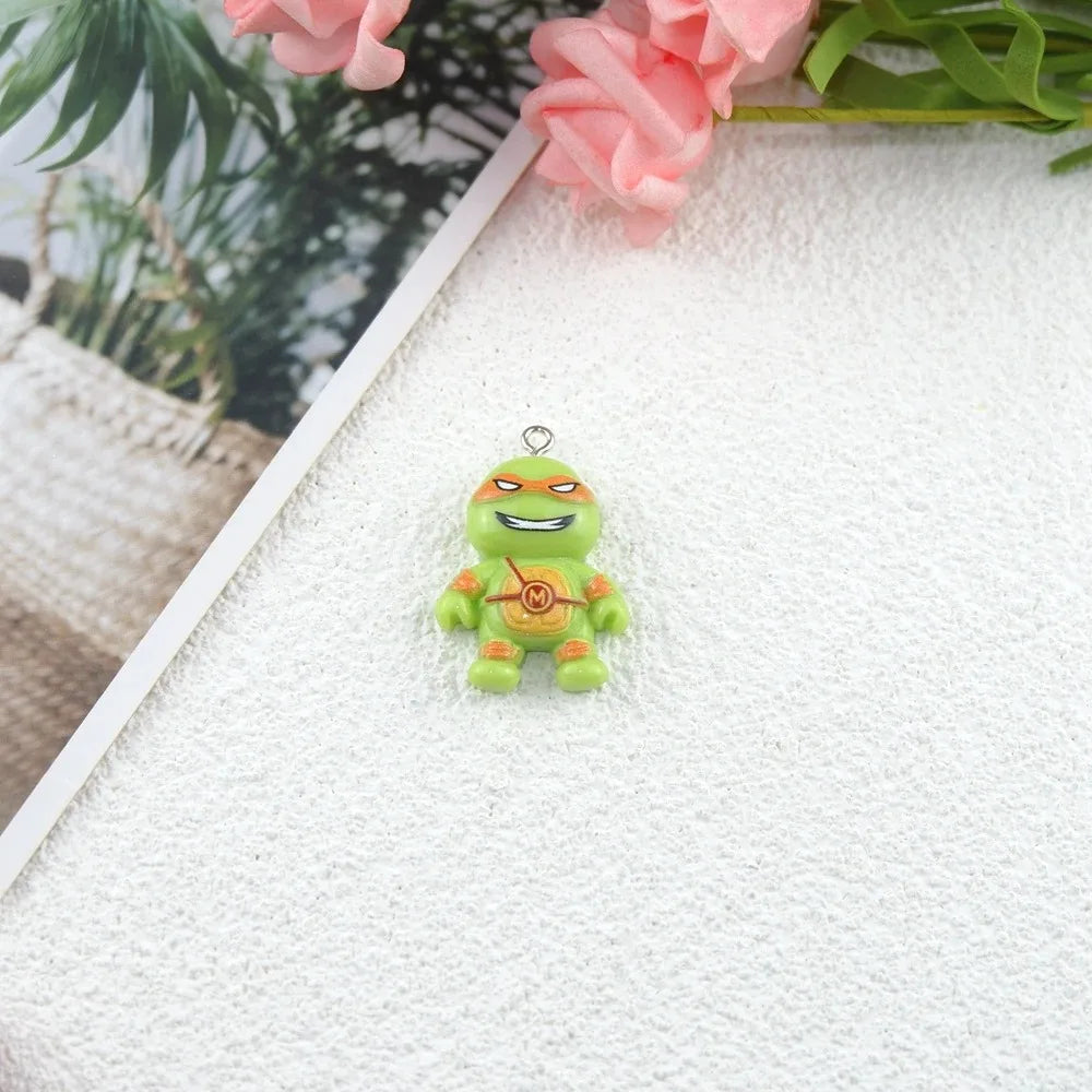 A small green cartoon turtle charm with orange accents and a metal loop at the top, placed on a textured white surface with pink flowers and green leaves in the background.