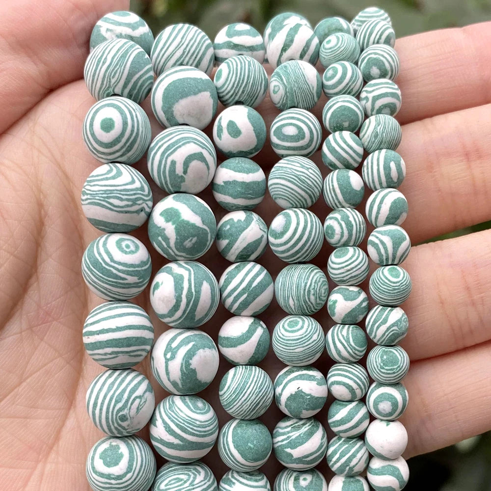 A close-up of multiple strands of round beads held in a person's hand. The beads have a marbled swirl pattern in sage green and white.