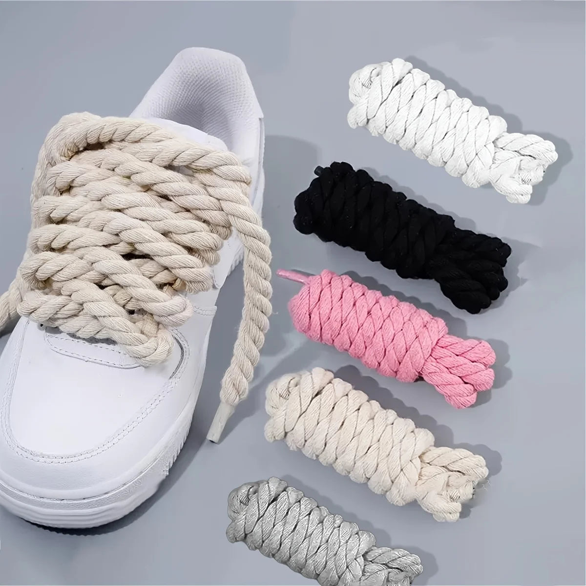 White sneaker with thick cream shoelaces, accompanied by additional shoelaces in black, pink, and gray on a gray background.