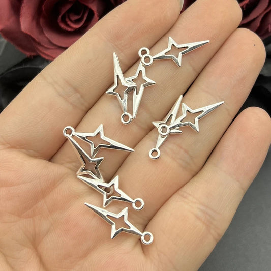 Five silver-toned metal charms shaped like stylized stars with small loops at the ends, resting on an open hand. The background includes blurred dark red roses and a dark surface.