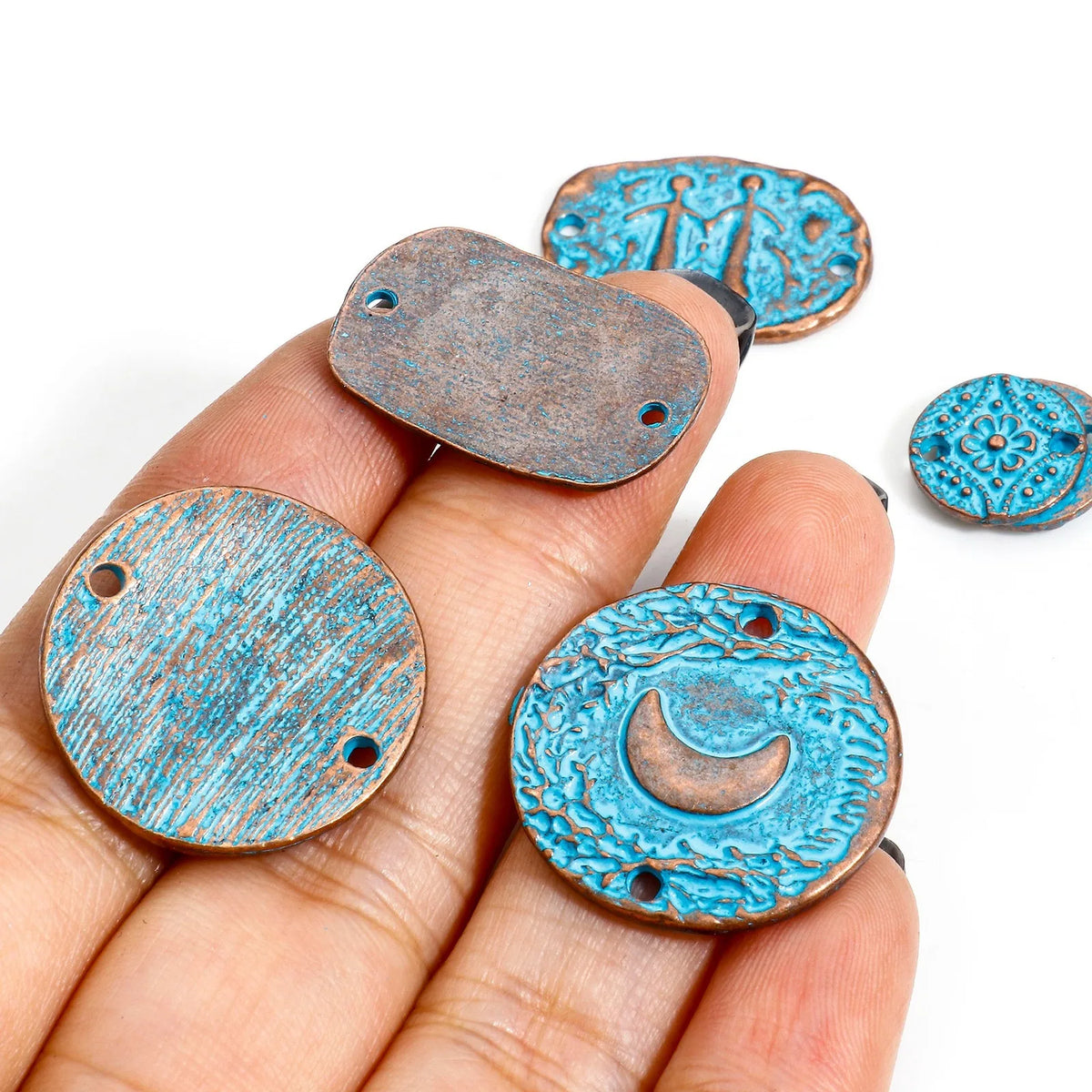 Five rustic turquoise-blue and bronze metal jewelry charms with screw holes are displayed on a person's hand against a white background.