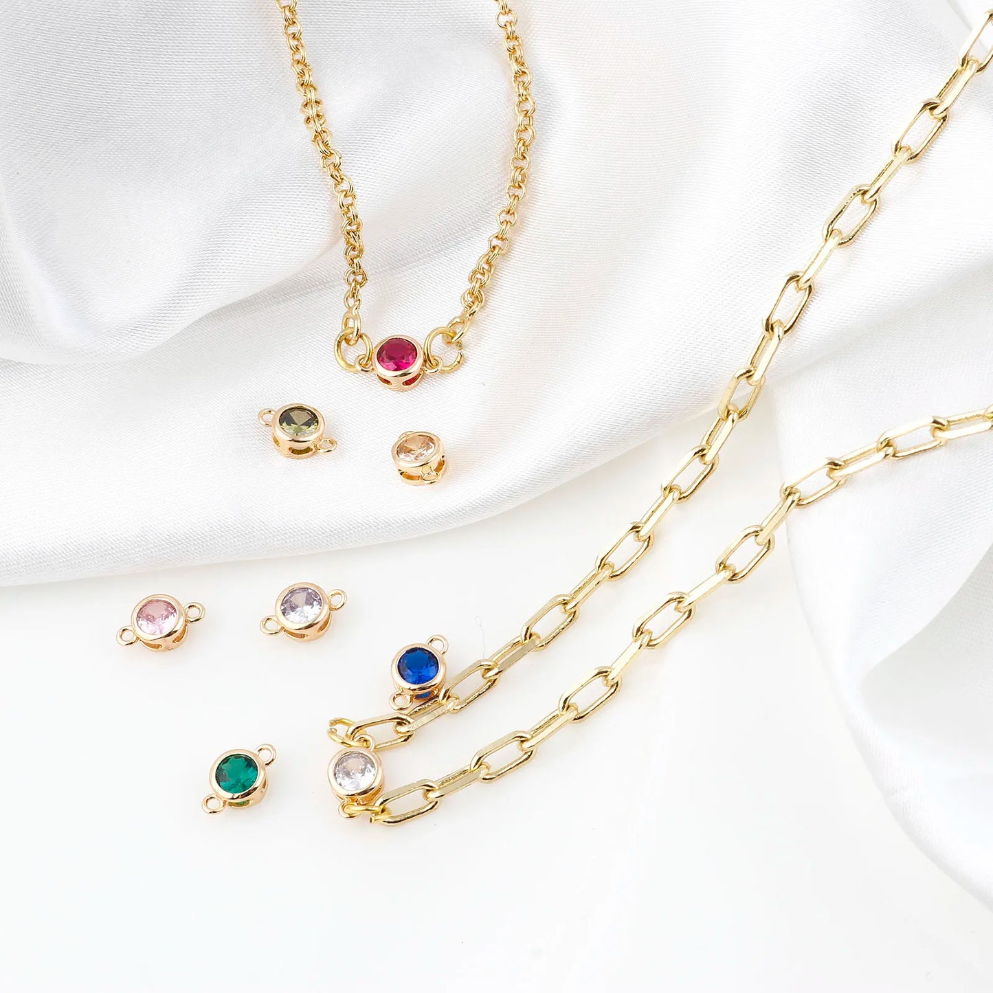 Gold chain jewelry set with a paperclip-style chain and small oval charm links, arranged on white draped satin fabric. The set includes several colorful gemstone charms in pink, green, blue, and clear settings.