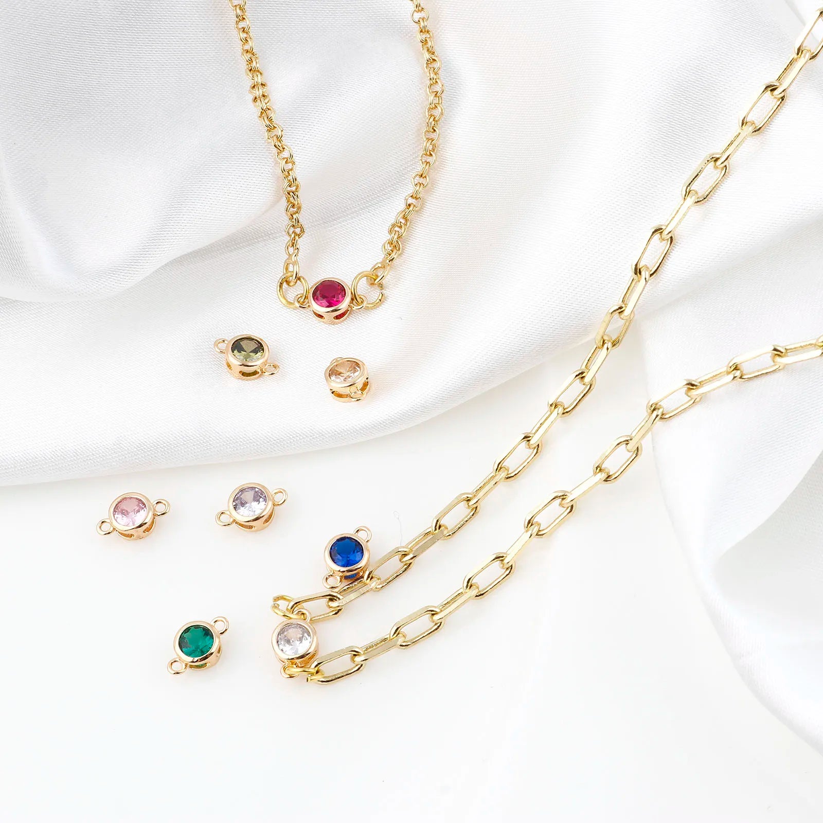 Gold chain jewelry set with a paperclip-style chain and small oval charm links, arranged on white draped satin fabric. The set includes several colorful gemstone charms in pink, green, blue, and clear settings.