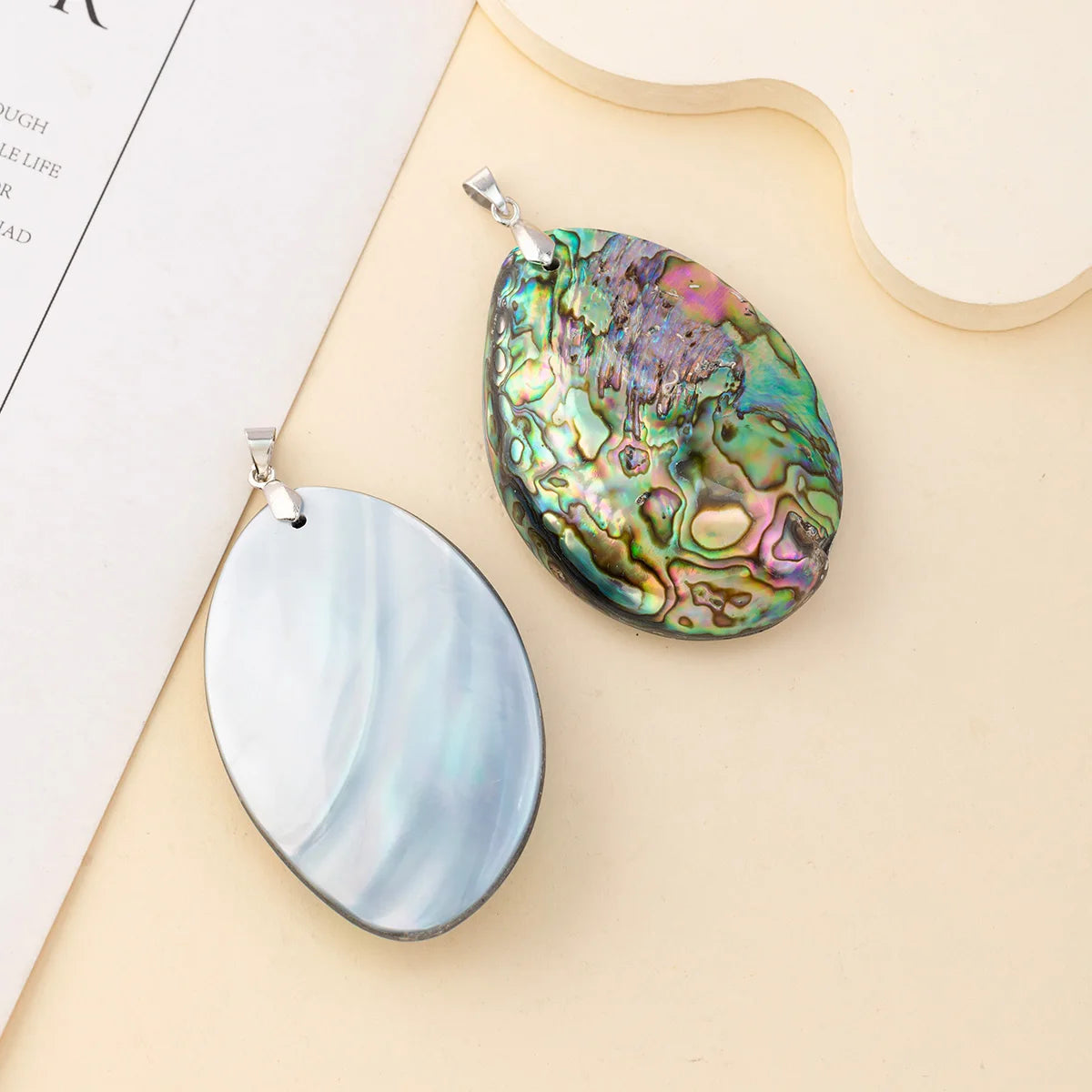 Two teardrop-shaped pendant charms displayed on a beige surface with a paper prop. One pendant has a smooth, iridescent blue-gray shell surface, and the other has a highly textured, shimmering abalone shell pattern with green, gold, and purple hues.