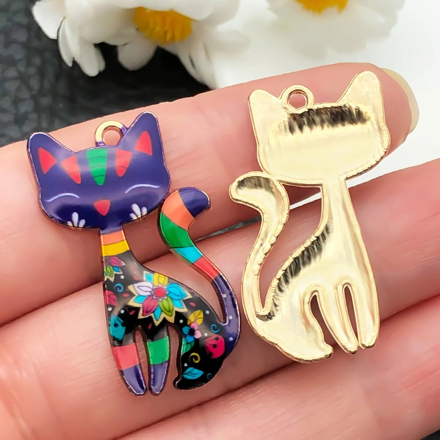 Colorful cat-shaped pendant and gold cat-shaped charm held between fingers with a blurred floral background.