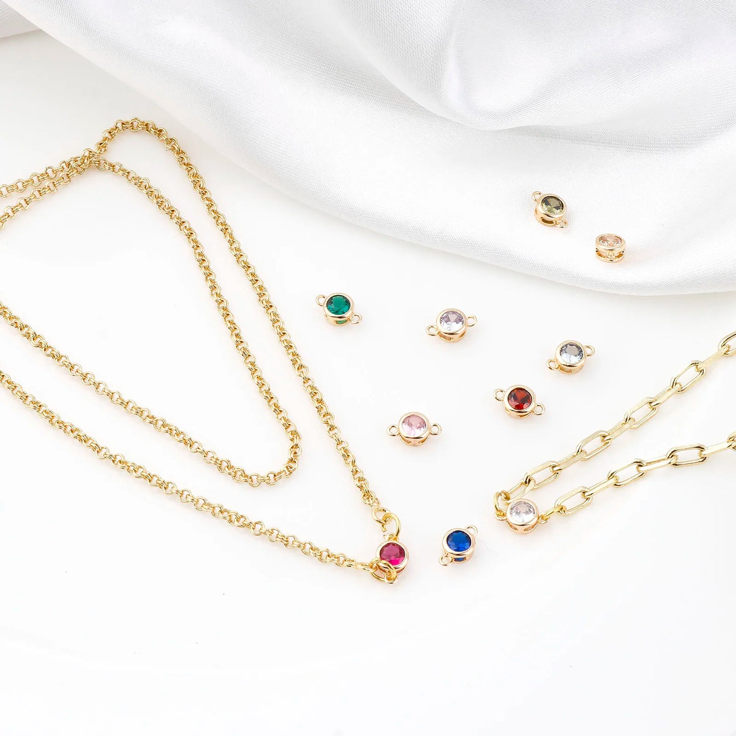 Gold chain necklaces and matching stud earrings with small round multicolor gemstone settings, arranged on a white fabric background.