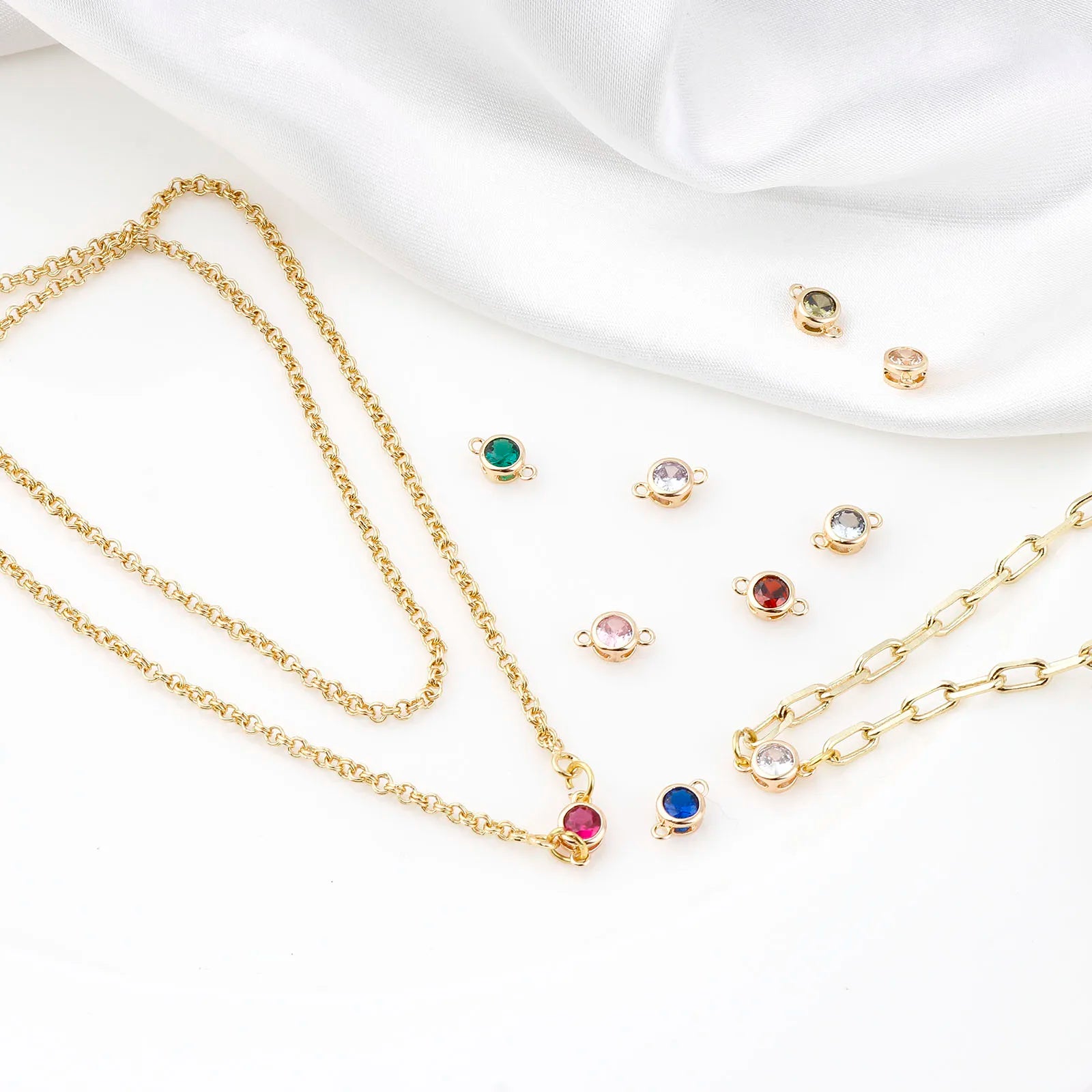 Gold chain necklaces and matching stud earrings with small round multicolor gemstone settings, arranged on a white fabric background.
