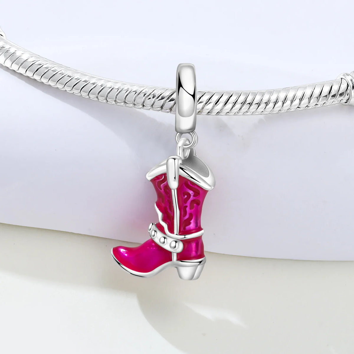 A silver-toned charm shaped like a cowboy boot with bright pink enamel detailing, shown hanging from a silver snake chain bracelet against a clean white background.