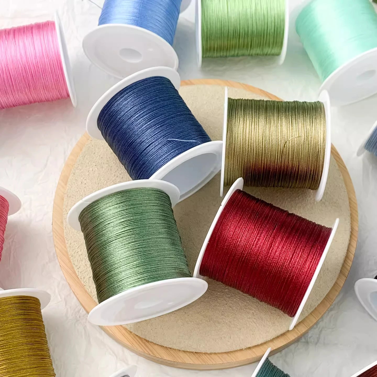 Multiple spools of thread or cord in assorted colors, including pink, blue, green, gold, and red, arranged on a round wooden tray against a light marble-like background.