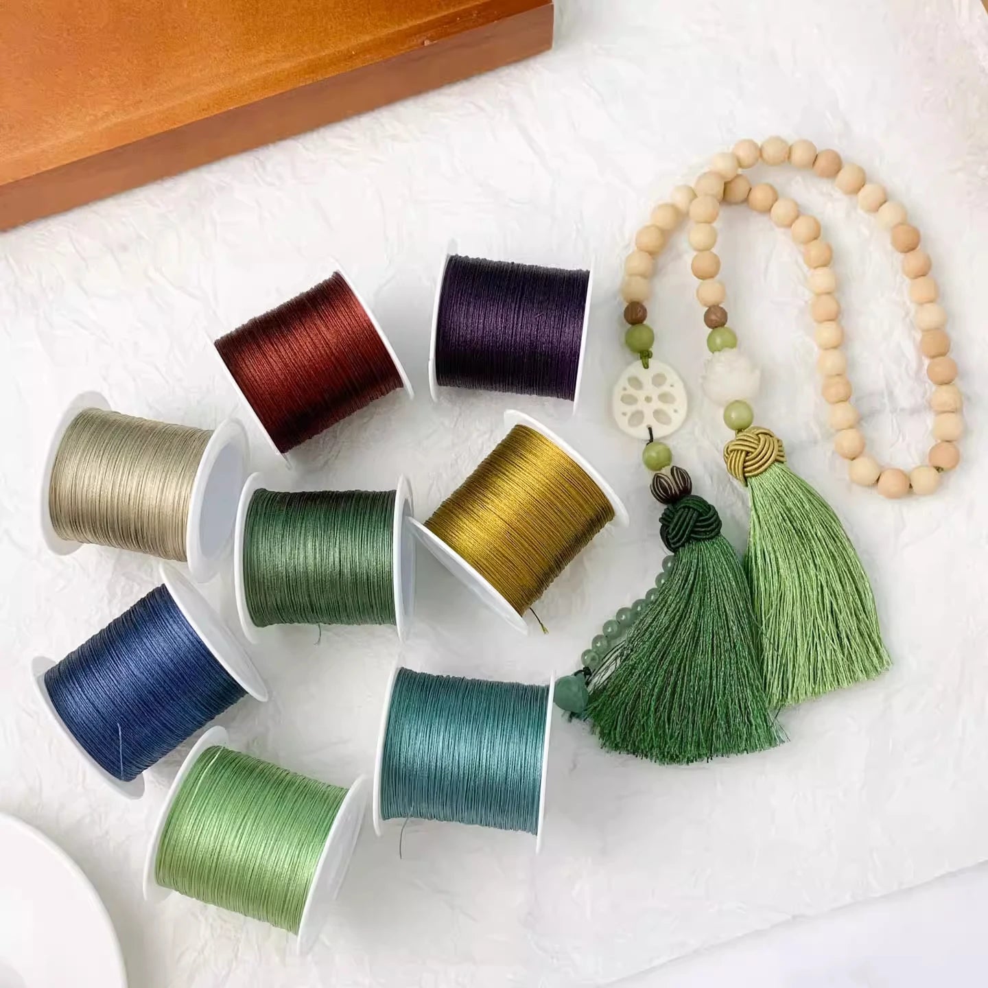 A styled flat-lay of colorful embroidery floss spools in shades of red, purple, beige, green, blue, and teal, alongside a wooden bead necklace with green tassels, arranged on a white textured fabric.