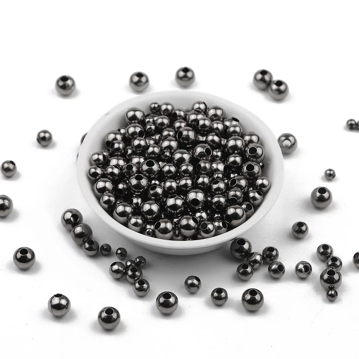 Small metallic dark gray beads with hollow centers, shown piled in a white shallow dish with a few scattered around on a white background.