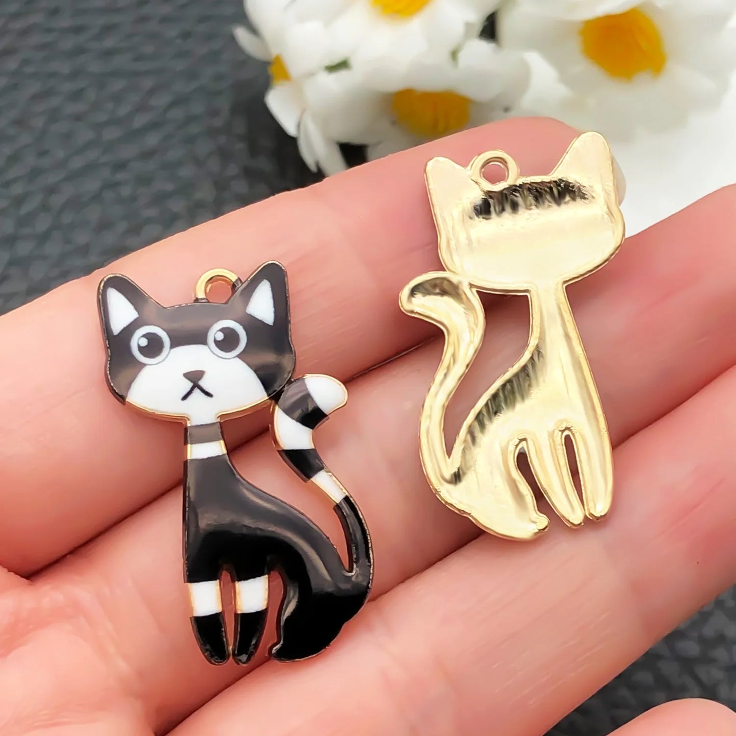 Two cat-shaped charms, one black and white, one gold, held between fingers with a blurred floral background.