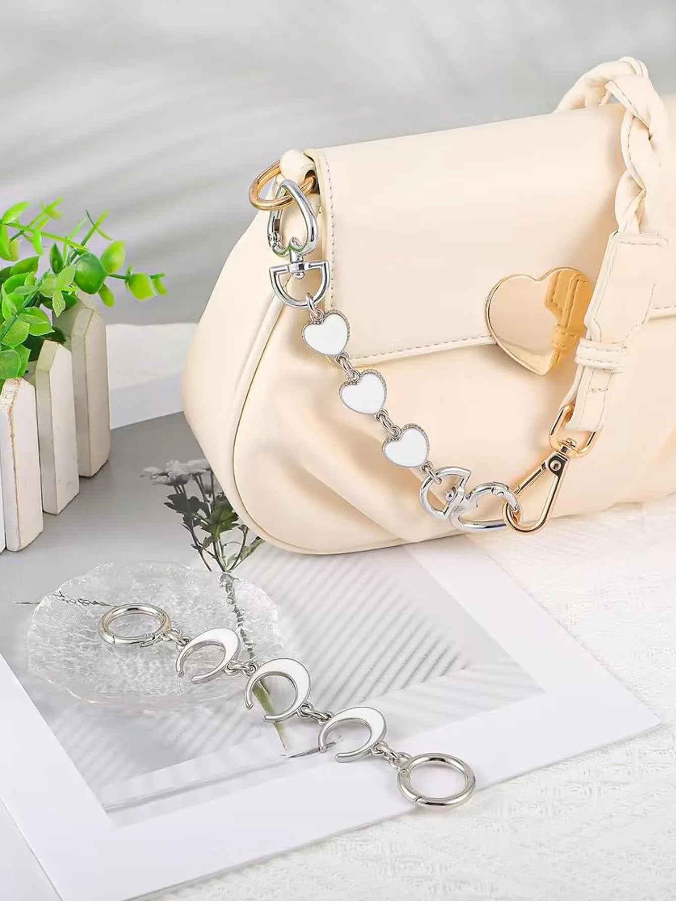 A cream-colored faux leather shoulder bag with a decorative silver chain strap featuring white heart-shaped links, displayed on a white surface next to a small potted plant.