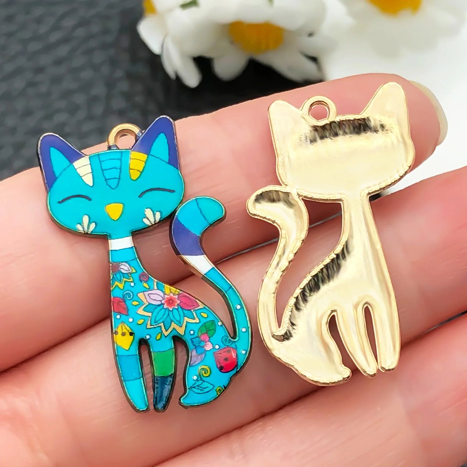 Colorful cat-shaped pendant and gold cat-shaped charm held between fingers with a blurred floral background.