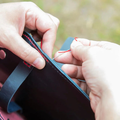 Close-up of hands stitching leather with red thread against a blurred natural background