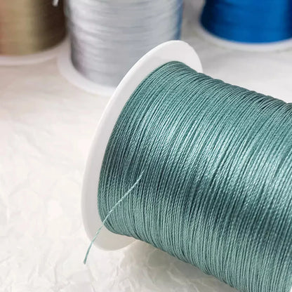 A close-up of a spool of mint green sewing thread, with other spools of thread in silver, gold, and blue blurred in the background on a white surface.