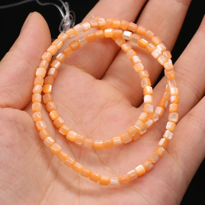 Orange and white faceted square shell beads strung on a cord, coiled and held in a person's hand against a black background.
