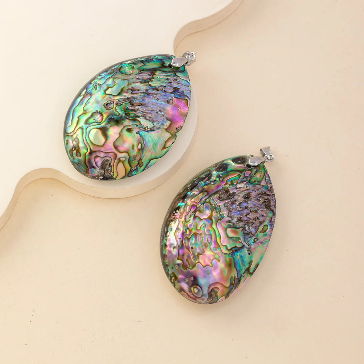 Two teardrop-shaped abalone shell jewelry pendants with silver-tone loops, displayed on a beige background with a wavy cream prop.