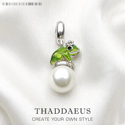 Jewelry pendant with pearl and green frog design on a white background, featuring the brand 'THADDAEUS'.