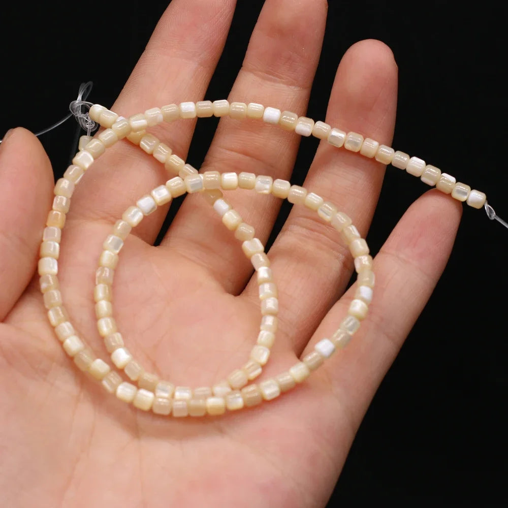 A hand holding a strand of small, cylindrical cream-colored shell beads with a glossy finish against a solid black background.