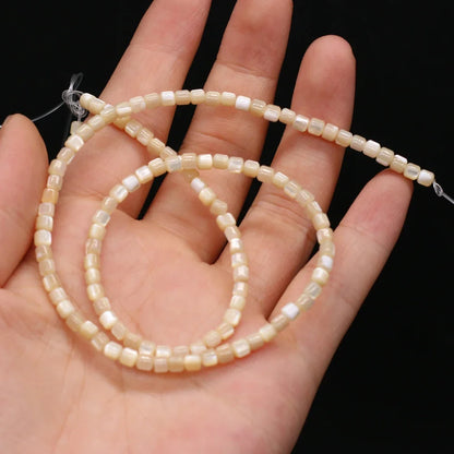 A hand holding a strand of small, cylindrical cream-colored shell beads with a glossy finish against a solid black background.