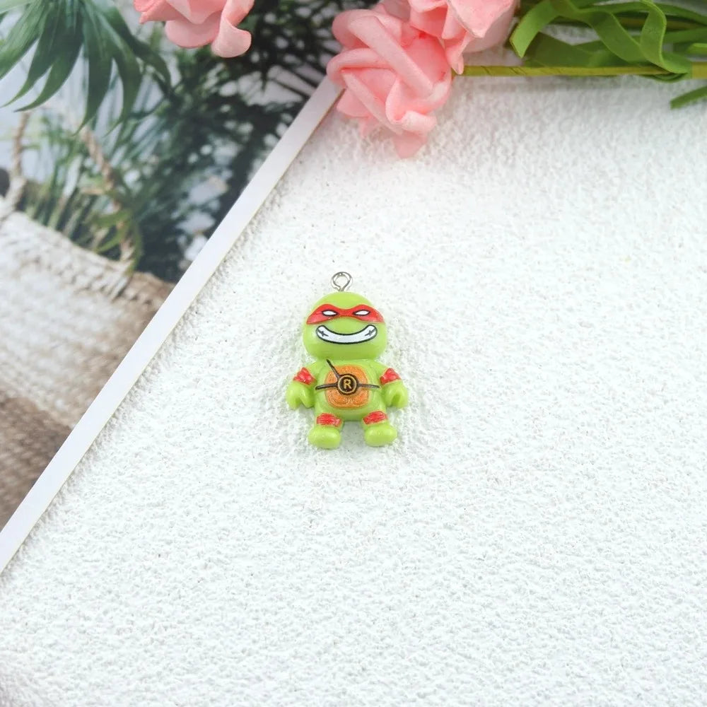 A small green cartoon character charm with red mask details and a silver hanging loop, placed on a textured white surface with pink flowers and a blurred plant in the background.
