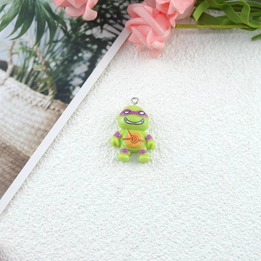 A small green cartoon turtle charm or pendant with purple headband and bracelets, standing on a textured white surface with pink flowers and blurred greenery in the background.
