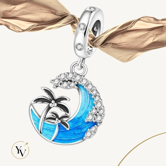 A silver charm bead featuring a crescent wave and palm tree design with blue enamel, clear rhinestones, and black palm tree details, shown hanging against a light beige background with a gold ribbon.