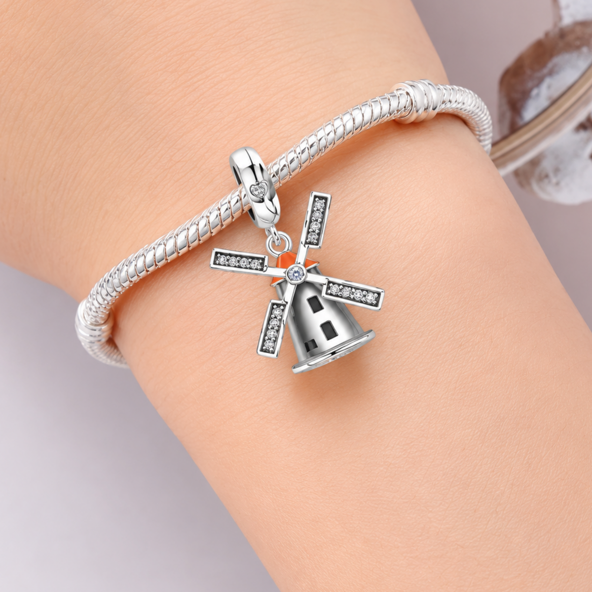 A silver snake bracelet with a dutch windmill charm, worn on a person's wrist. The windmill charm features a silver body with black and white striped blades, a small orange accent, and clear rhinestone details.
