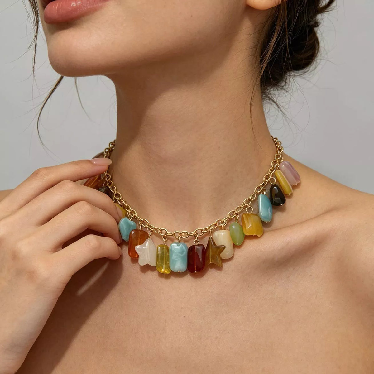 A close-up of a model wearing a gold chain necklace adorned with colorful, irregularly shaped stone charms in shades of blue, green, yellow, orange, red, and white.